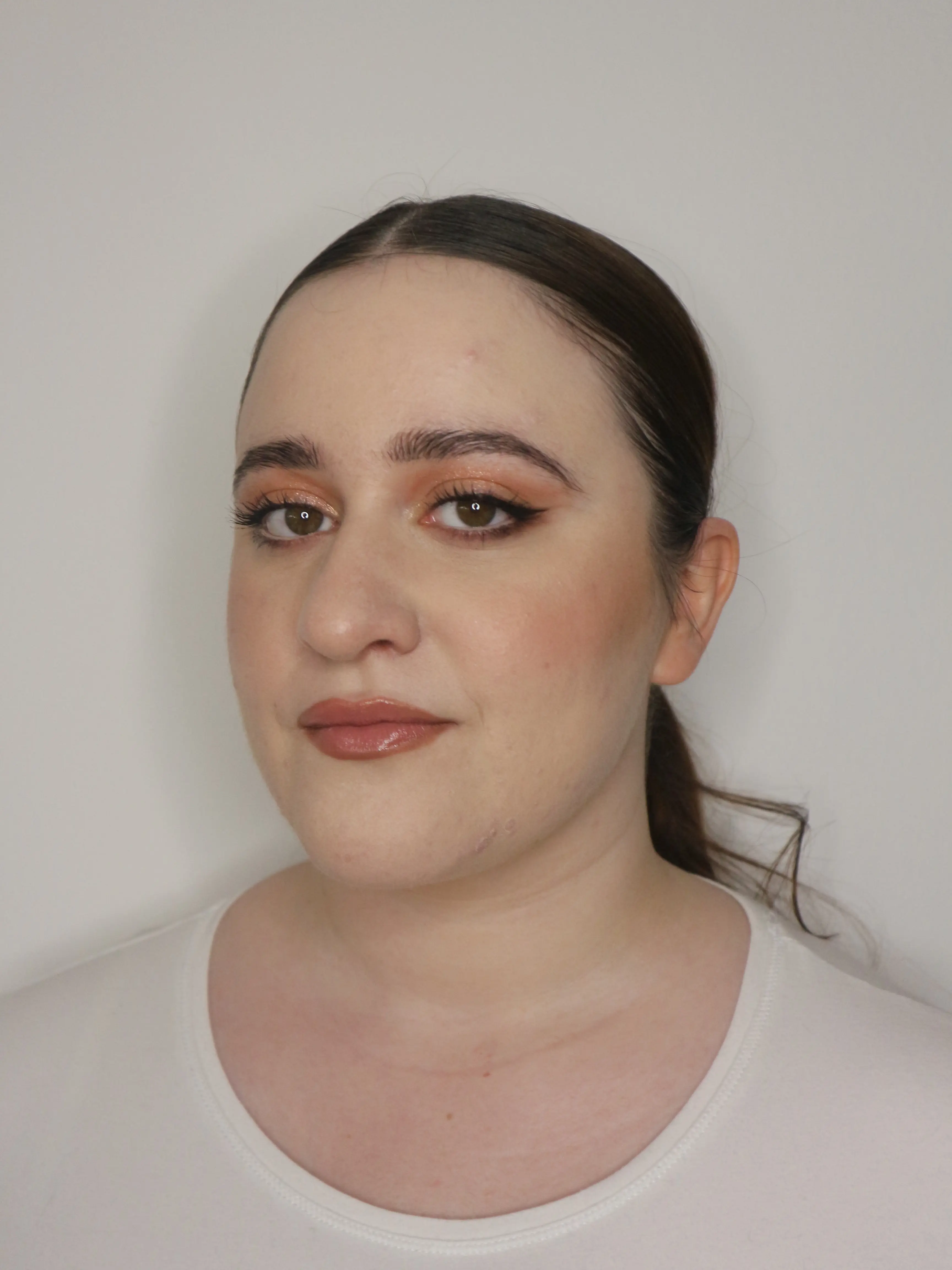 A radiant makeup look featuring coppery shades on the eyes, a graphic eyeliner that defines the gaze, and nude glossy lips for a natural yet refined elegance.